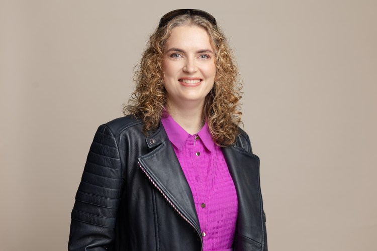Anica Zeyen is a white woman with curly dark blonde hair. she wears a purple pink dress with a black leather jacket. she smiles broadly