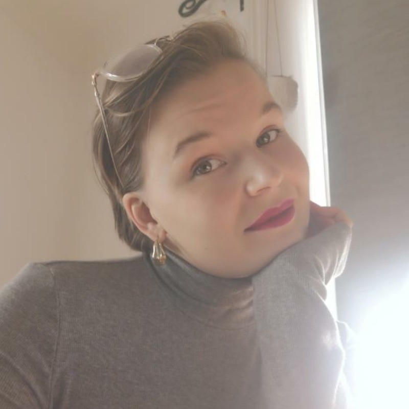 Headshot of Marianna Tarvainen. Softly lit portrait of a person with short hair and glasses resting on their head, wearing a grey turtleneck and gold earrings, looking calmly toward the camera.