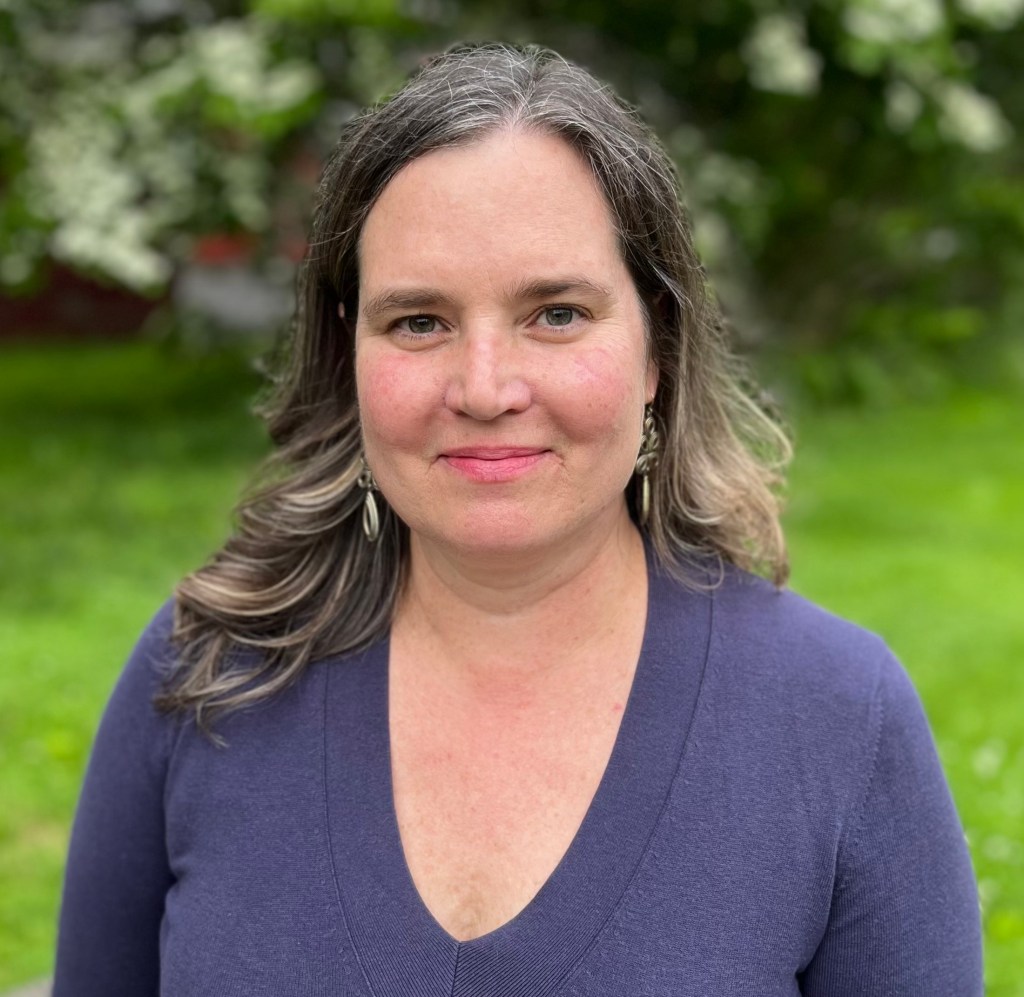 A white woman with wavy brown hair smiles at the camera. She is sitting outside, with green grass in the background. 