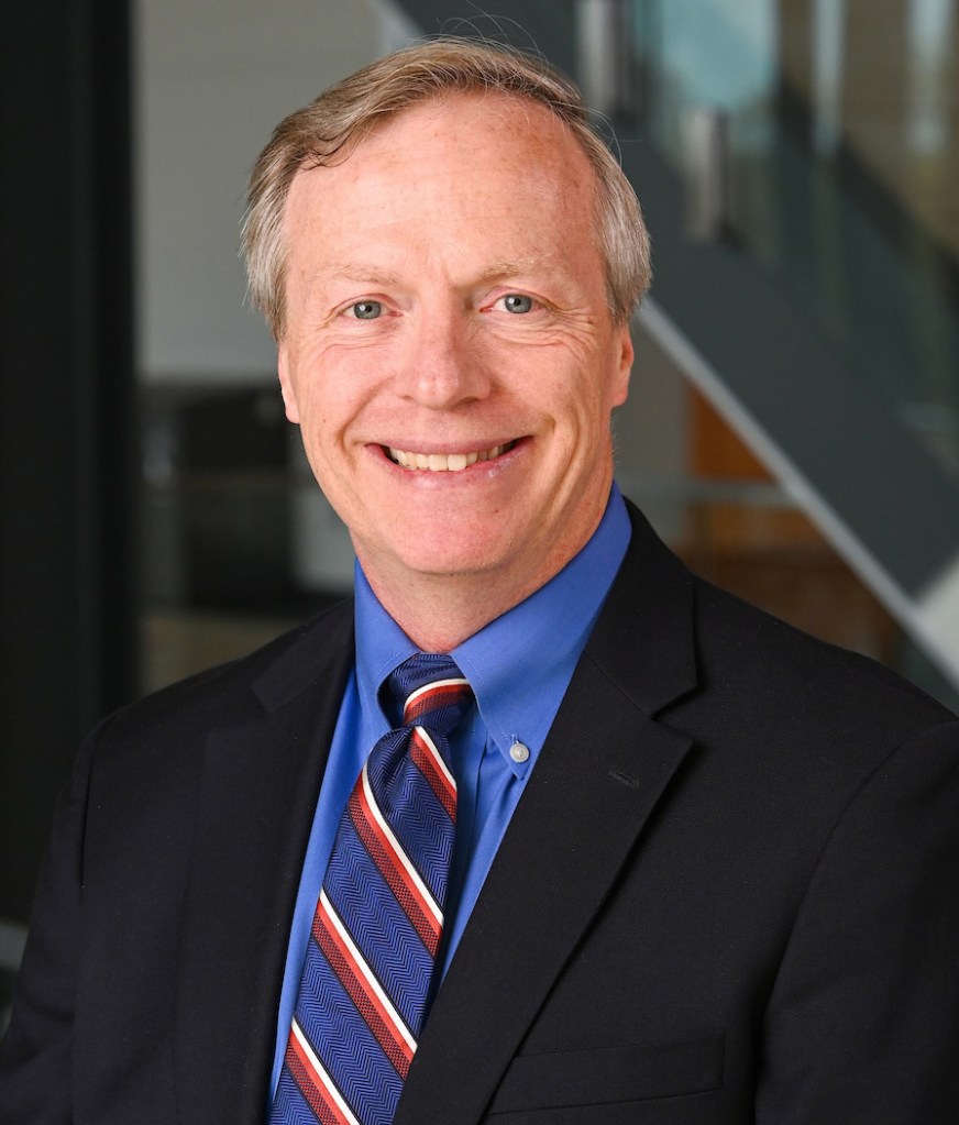  professional headshot of a smiling white male, with a black suit, blue dress shirt, and an orange and blue striped tie.