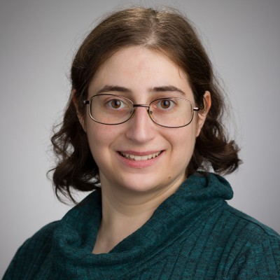 Headshot of Lauren Gilbert. She is a white woman with dark brown hair that is largely straight but curls near the ends. She wears glasses and a green sweater.
