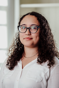 Headshot of Kyra Rodrigues. She is a mixed race Puerto Rican woman with brown curly hair. She wears a white button down, black glasses, and gold septum, nose, and necklace jewlery. The background is a blurred white room with a window.
