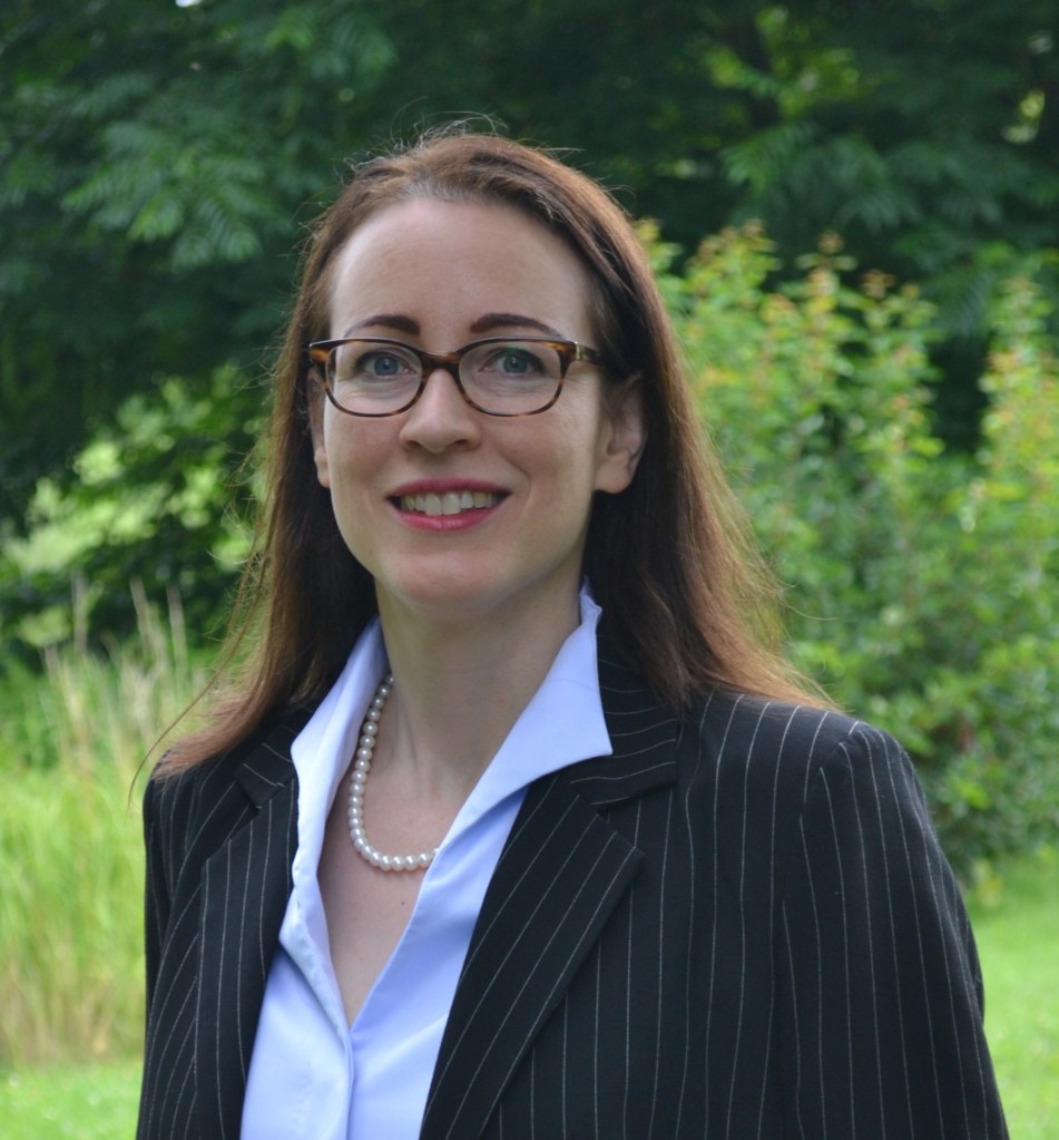 necklace, long brown hair and brown glasses) in the park of Trier University with bushes and trees in various shades of green in the background