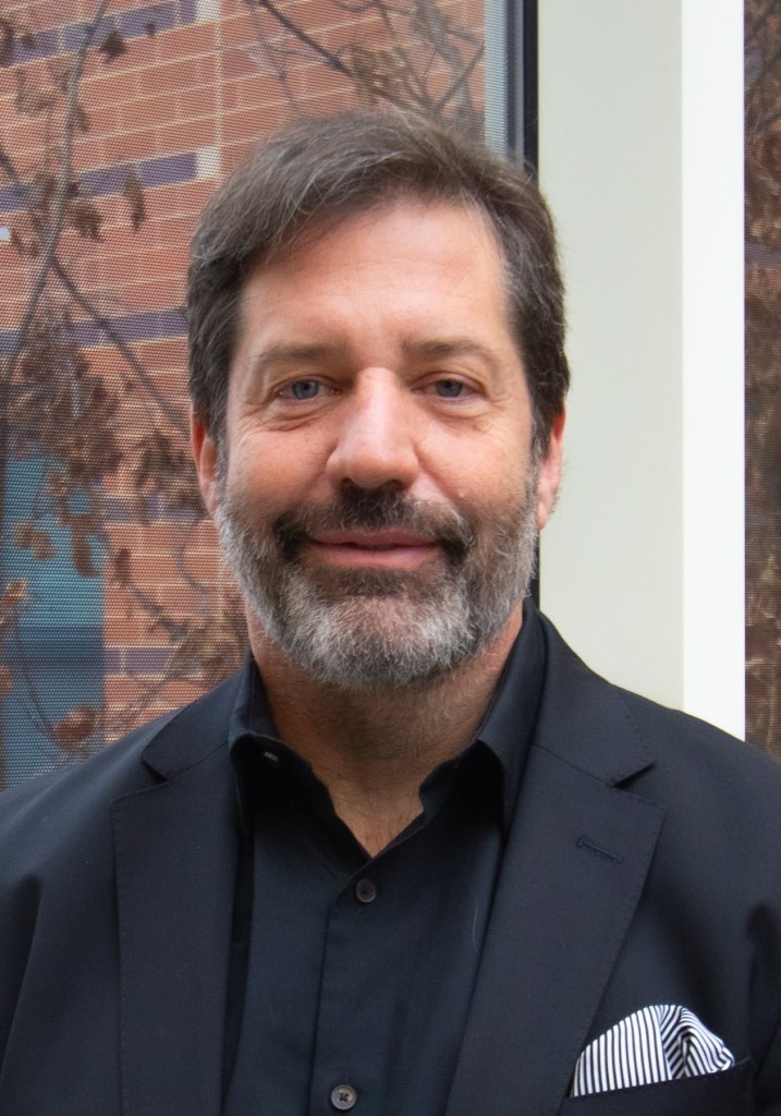 Informal headshot of Matt wearing a black blazer and a black top. He has a full beard . His hair is dark. 