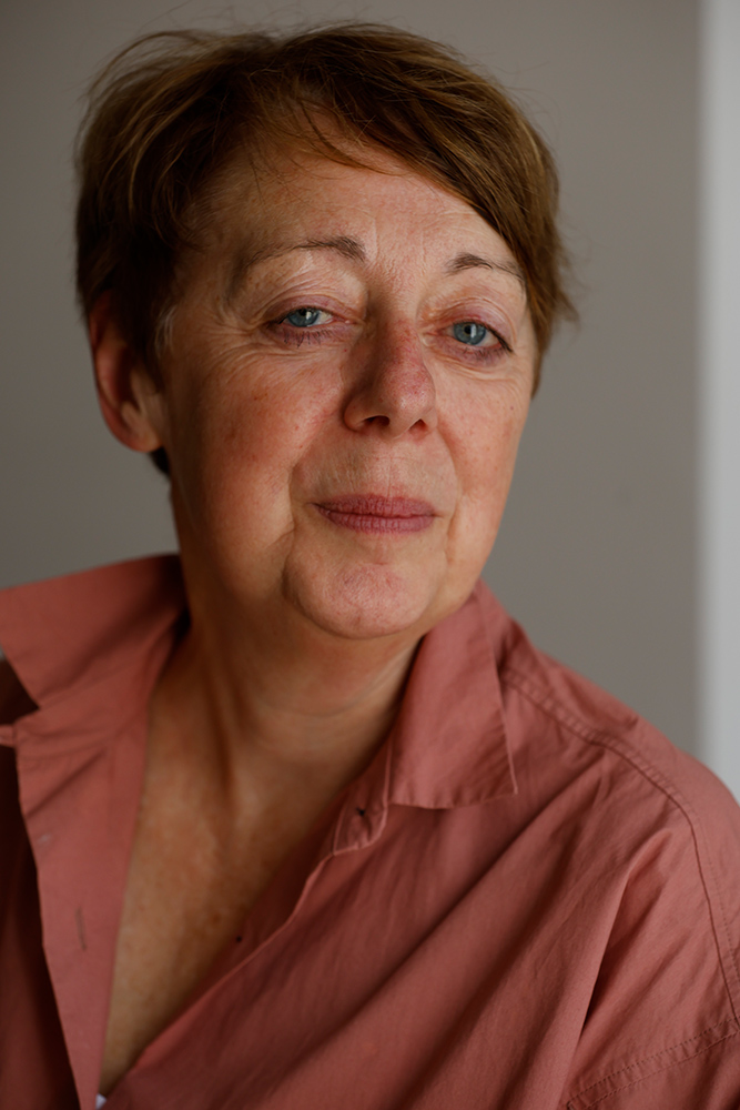Professional, close-up portrait of a Caucasian woman with short brown hair and light skin. She is wearing a muted rose-colored shirt and looks slightly downward with a calm, thoughtful expression against a plain, softly lit background.