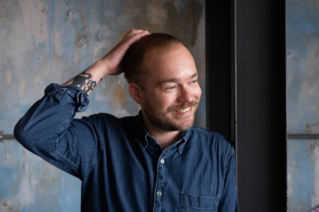 A man wearing a blue denim shirt stands indoors near a window, with one hand resting on the back of his head. A tattoo is visible on his forearm, and a textured wall is in the background.
